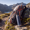 A drink bottle in blue metal, resting beside a backpack on rocky terrain, with mountains in the background.