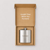 A stainless steel hip flask inside a plain brown box with a logo and light text on the interior.