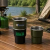 A stainless steel camping mug set, featuring a black, green, and metallic finish, displayed on a wooden table by a lake.