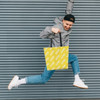 A person jumps while holding a vibrant yellow tote bag featuring a logo, set against a striped grey background.