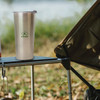 A drink bottle in stainless steel sits on a campsite table, with a logo on its side and a blurred background of nature.