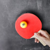 A table tennis paddle with a yellow ball resting on it, set against a dark background. The paddle features a logo.