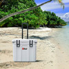 A white cooler box with wheels stands on a sandy beach surrounded by greenery and water. It has a logo.
