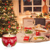 A red tin candle with a snowman design, placed near a festive dinner setting and Christmas tree.