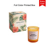 A scented candle in an orange glass container, accompanied by a colourful printed box featuring floral designs.