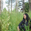 A unisex soft shell jacket in black, worn by a person sitting in a grassy area surrounded by trees.