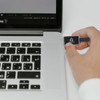 A black flash drive with a logo is being inserted into a laptop’s USB port. The laptop features a silver design.
