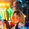 A person with curly hair wearing pink sunglasses and a colourful outfit, raising their arm in a lively outdoor event setting.