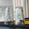 A set of clear glass tumblers, one featuring a design with text and a logo, the other is plain.