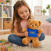 A smiling girl holds a light brown bear plush toy wearing a blue shirt. The background features colourful play items and a cozy setting.