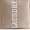 A laundry basket in a beige fabric with white lettering, featuring a sturdy handle and a simple design.