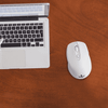 A white wireless mouse placed next to a silver laptop on a wooden desk. The laptop has a visible keyboard.