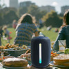 A black outdoor Bluetooth speaker with an LED light strip, set on a picnic table amidst food and drinks.