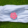 A pink soft enamel golf ball marker featuring a floral design, placed on a white golf glove on green grass.