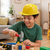 A child wearing a bright yellow PVC construction hat smiles while using a hammer on a wooden building toy.