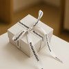 A white gift box tied with a patterned satin ribbon featuring a logo. The ribbon is primarily white with black text.