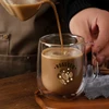 A barrel-shaped double wall mug in clear glass is being filled with a creamy coffee beverage.