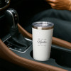 A drink bottle in a car cup holder, featuring a white recycled stainless steel design with a clear lid and a logo.