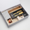 A gift box containing a wooden cheese board, a gold cheese knife, a jar of chutney, and a striped cloth.