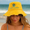 A yellow bucket hat with a frayed edge and a logo embroidered on the front, worn by a person at the beach.
