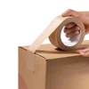 A hand holds a roll of brown self-adhesive kraft paper tape, partially applied to a cardboard box.