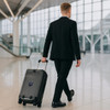 A black hardside suitcase on wheels is being pulled by a man in a suit in an airport terminal.