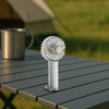 A transparent fan with LED light, featuring a white handle, on a table outdoors beside a coffee mug.