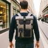 A roll-top backpack in grey and black, featuring multiple pockets, being carried by a person on a street.