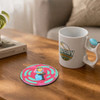 A colourful PU coaster featuring a cartoon design, placed beside a mug on a wooden table.