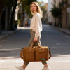 A woman carrying a foldable travel duffle bag in brown while walking on a street. The bag features a logo.