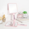 A pink handheld square mirror with a handle, placed alongside perfumes and decorative items on a surface.