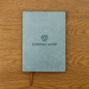 A soft PU leather notebook in a muted green colour with a logo on the cover, placed on a wooden surface.