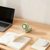 A foldable handheld fan in mint green placed on a wooden desk alongside a laptop and open books.