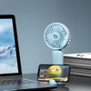 A light blue handheld fan with a phone stand, positioned next to a laptop and stacks of magazines.