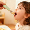 A green silicone and stainless steel spoon is being offered to a young child. The child appears eager to eat.