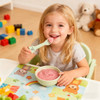 A child smiles while holding bear-shaped green silicone cutlery, enjoying a bowl of pink yoghurt. Colourful toys are in the background.
