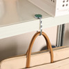 A round foldable desk hook in silver, attached to a shelf, with a tan handbag hanging from it.