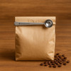 A brown paper bag of coffee with a stainless steel measuring spoon resting on top, surrounded by coffee beans.