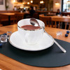 A white cup with a gold rim sits on a saucer, complemented by a silver spoon and scattered blueberries.