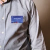 A person wearing a checkered shirt with a blue custom PVC name badge attached. The badge features a logo.
