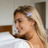 A wavy rectangle iridescent hair clip in light purple is styled in long, blonde hair. A simple room is in the background.