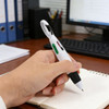 A hand holding a white and black carabiner pen over an open notebook, with a computer and binders in the background.