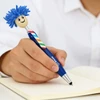 A blue pen with a smiling character head and colourful striped body, being held while writing in a notebook.