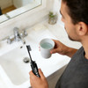 A black foldable toothbrush held by a person next to a sink, with a cup of water in the other hand.
