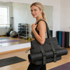 A black tote bag with yoga mat straps is being carried by a person in a fitness studio. The bag has a logo.
