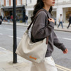 A beige canvas tote bag with brown straps, carried by a person in a grey sweater while walking on a city street.