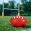 A red mini promotional Aussie Rules ball resting on green grass, with goalposts blurred in the background.