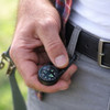 A black carabiner compass thermometer hook is held in a hand, attached to a belt, against a blurred outdoor background.