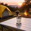 A compact stainless steel folding cup rests on a table, with a tent, stove, and campfire in the background.