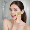 A flower-shaped facial cleansing brush in gold is being held against a woman's cheek. The setting is a bathroom.